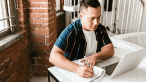A focused adult student sitting at a desk in a bright room, writing notes in a notebook while looking at an open laptop—symbolising flexible learning and mature study.