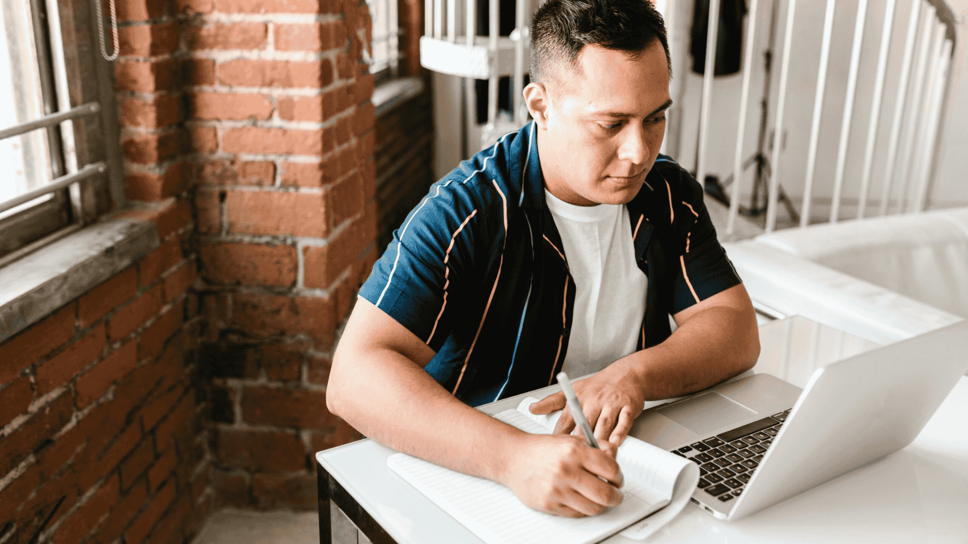 A focused adult student sitting at a desk in a bright room, writing notes in a notebook while looking at an open laptop—symbolising flexible learning and mature study.