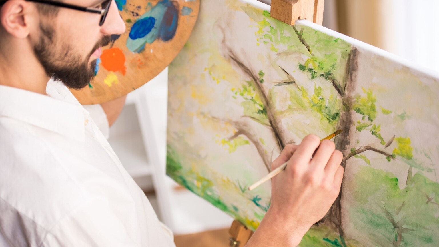 Online creativity and arts courses from NEC. Male paiting a tree on canvas holding a wooden palette of paint.