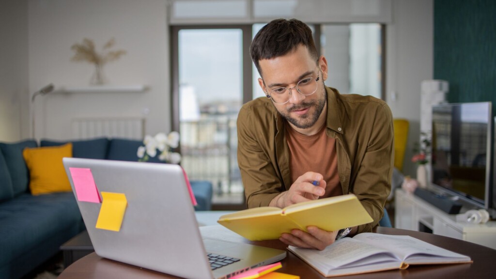 Online develop your skills. Male looking at a book while using a laptop sat at a desk.