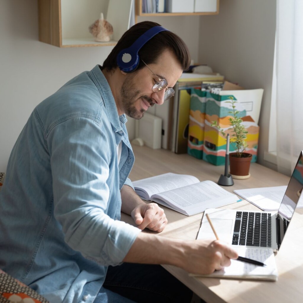 Online teaching courses with the NEC, . Male with headphones working on a laptop sat at a desk in front of a window, taking notes i a note book with a yellow pencil.