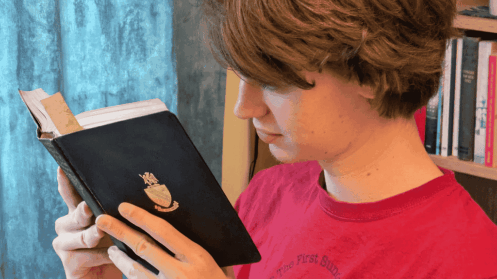 A young student in a red T-shirt is deeply focused on reading a black hardback book with a crest on the front. He stands in front of a blue curtain, beside a bookshelf filled with books. The scene reflects concentration, curiosity, and a love for literature.
