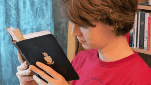 A young student in a red T-shirt is deeply focused on reading a black hardback book with a crest on the front. He stands in front of a blue curtain, beside a bookshelf filled with books. The scene reflects concentration, curiosity, and a love for literature.