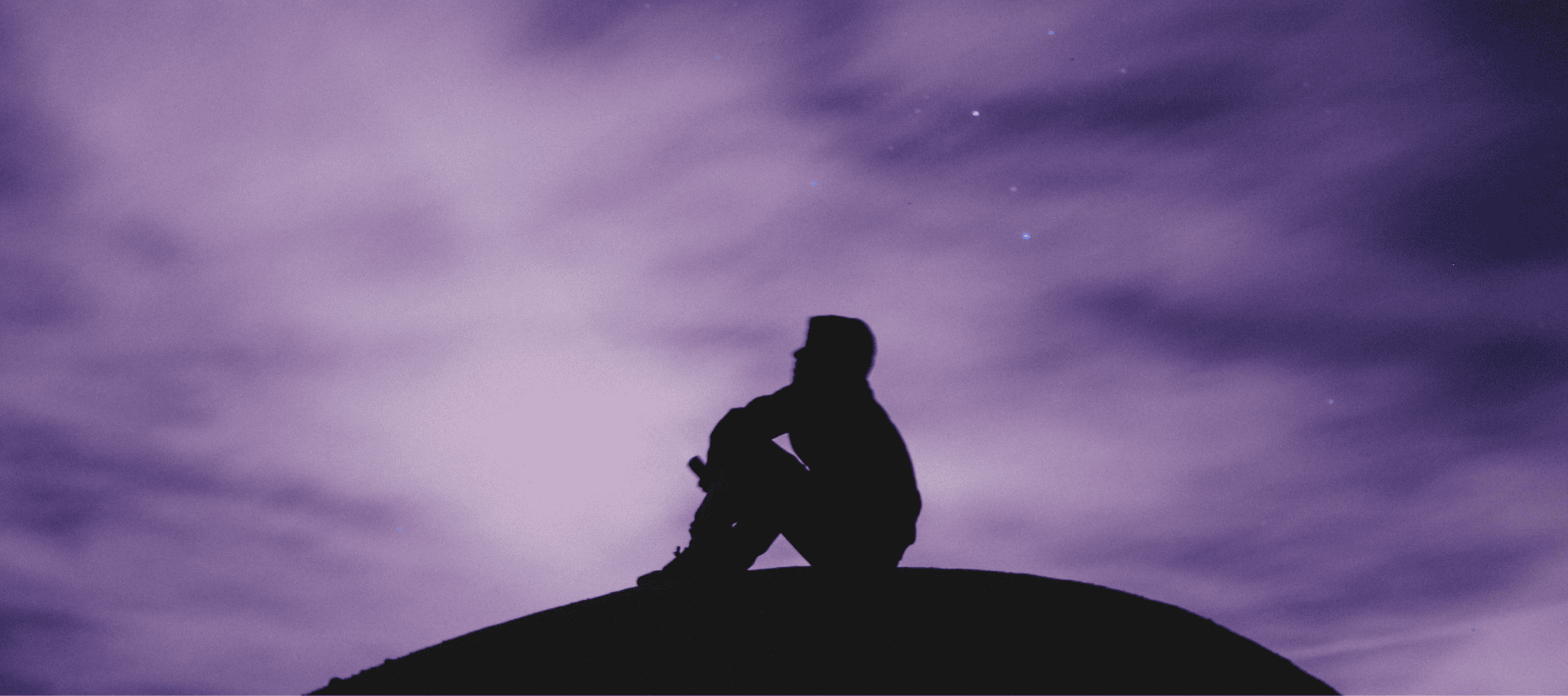 A man sits atop a hill at twilight, thinking