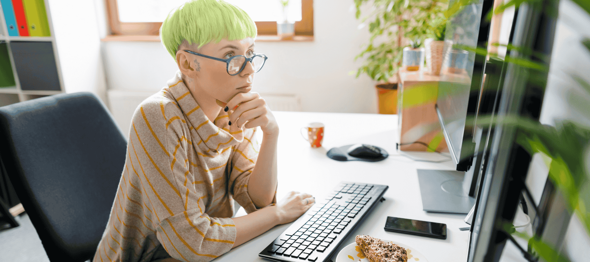 A girl sits at her desk in a sunny room at home concentrating on the big screen in front of her.