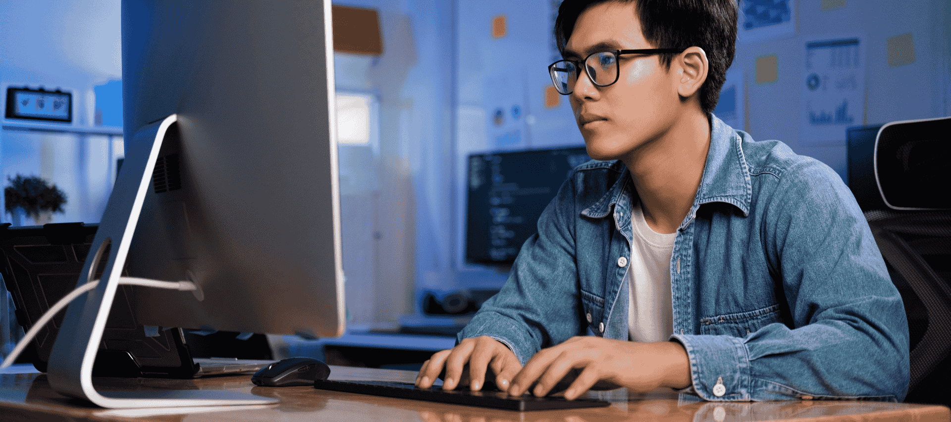 Teenager sits in a room using a computer with lots of other computers showing computer script around him