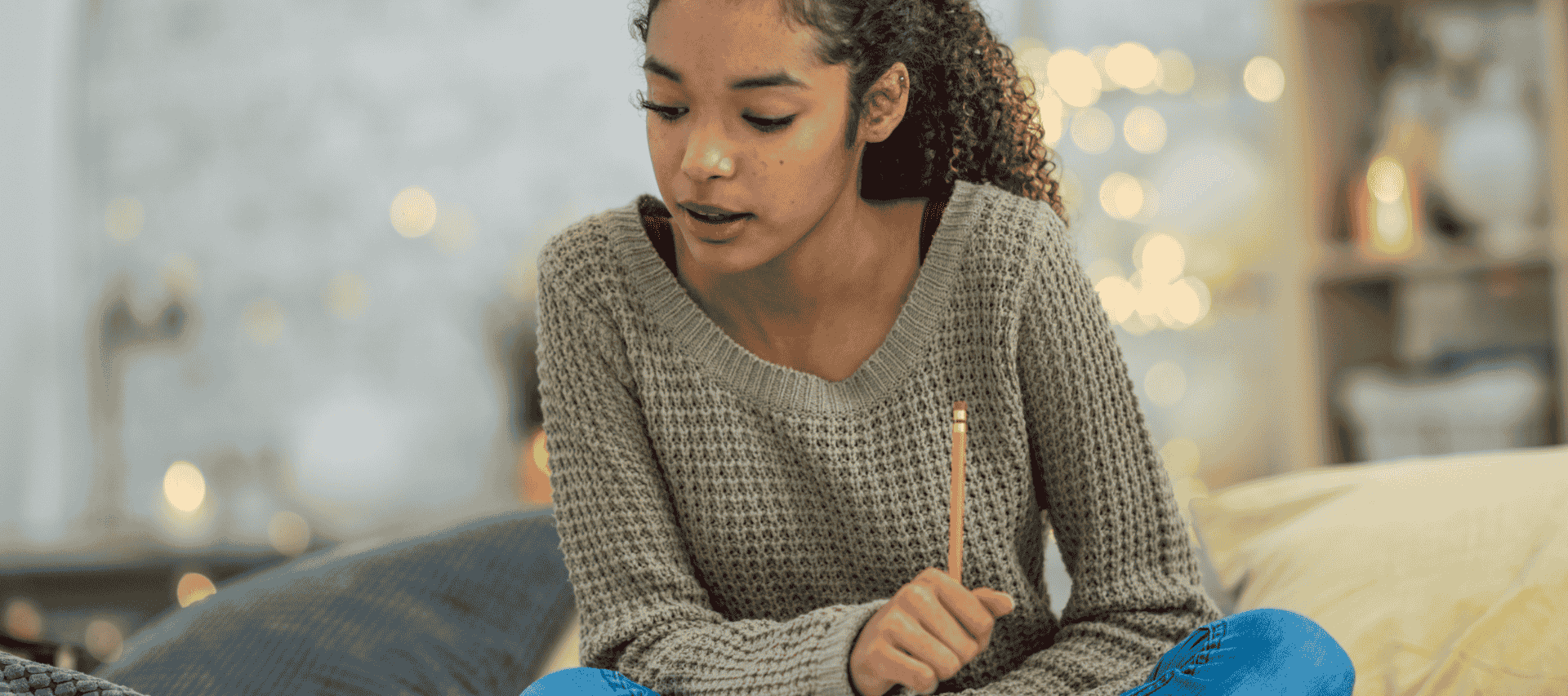 A teenage girl sits cross-legged on a sofa at home studying her course with pretty twinkling lights in the background.