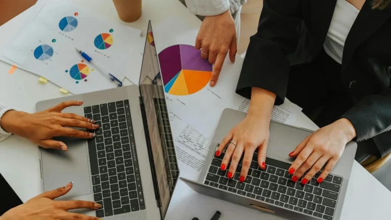 Person typing on a laptop with financial reports, spreadsheets, and a pie chart on the desk—suggesting business planning or financial analysis in a work or study setting.