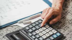 A close-up of a person using a calculator with worksheets and pens on a carpeted surface, representing focused mathematical problem-solving.