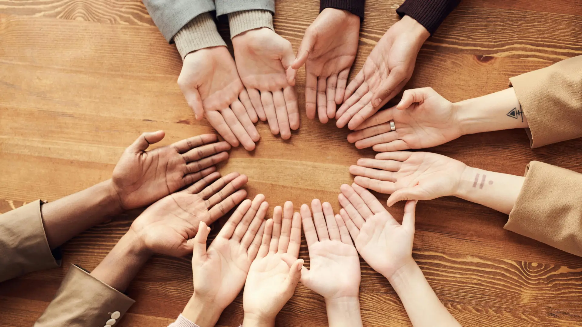 A group of diverse hands arranged in a circle on a wooden table, symbolising unity, inclusion, and social connection—key themes in sociology.