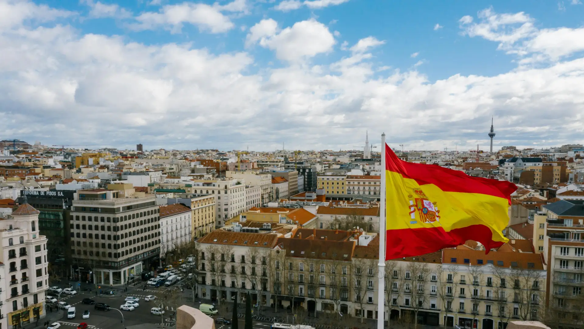 The Spanish flag waves above a cityscape in Madrid on a partly cloudy day, symbolising language, culture, and national identity