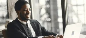 A smiling professional in a suit works on a laptop at a modern café, with a coffee beside him—reflecting career progression, flexibility, and success through distance learning CMI management qualification.