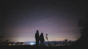 Two people star gazing looking over a town with telescope next to them. The night sky is covered in stars