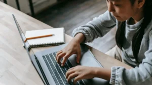 A focused student typing on a laptop at a desk with a notebook and pencil nearby—illustrating independent online learning at home.