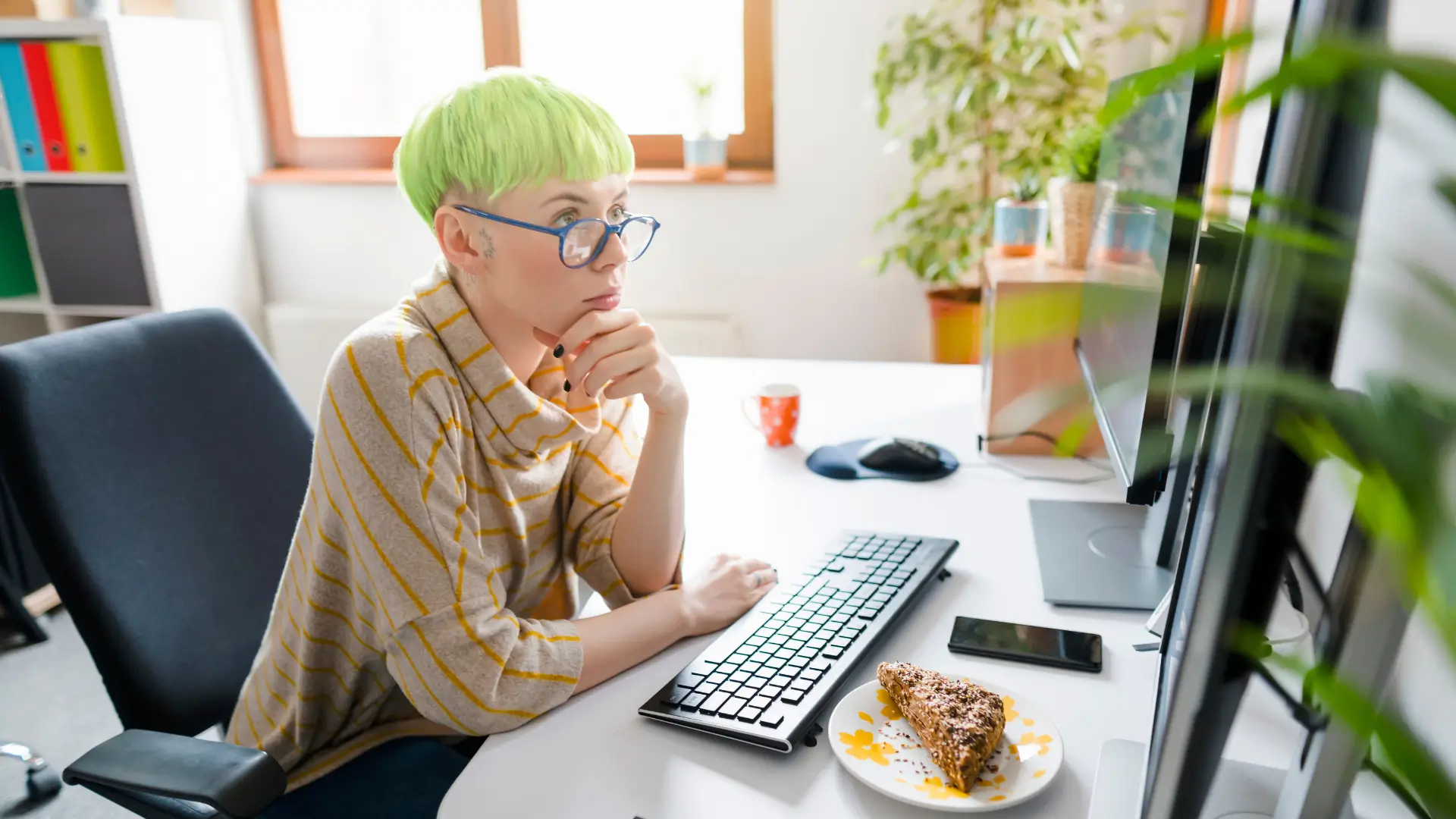 A girl sits at her desk in a sunny room at home concentrating on the big screen in front of her.