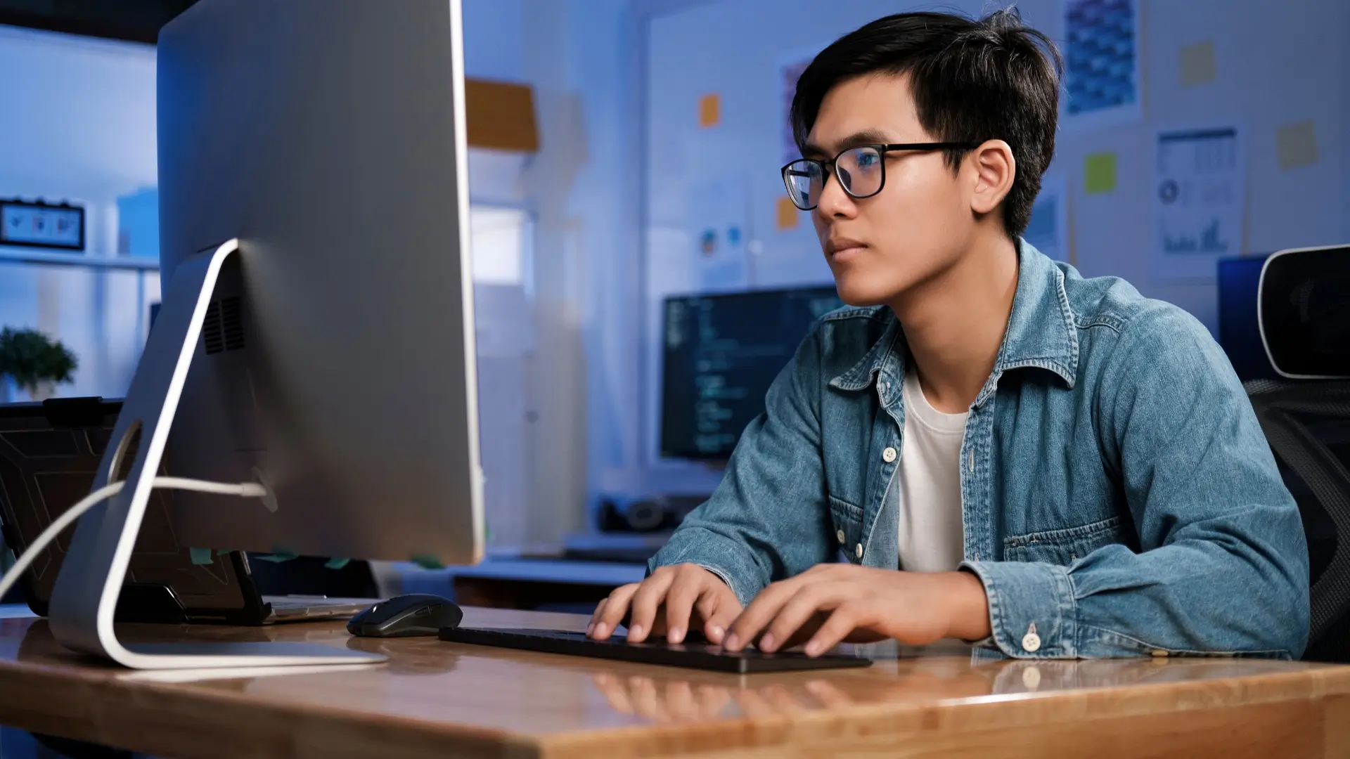 Teenager sits in a room using a computer with lots of other computers showing computer script around him