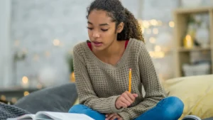 A teenage girl sits cross-legged on a sofa at home studying her course with pretty twinkling lights in the background.