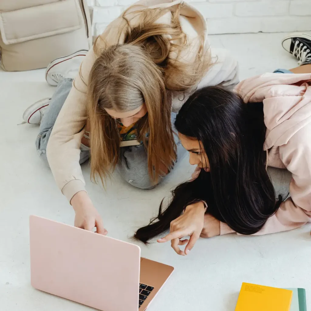 Two teenage girls in casual dress crouched on the floor studying and looking at information on a pink laptop