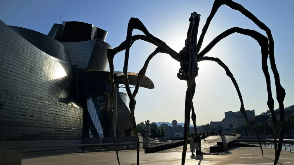 Louise Bourgeois’s giant spider sculpture Maman (1999) outside the Guggenheim Museum Bilbao, silhouetted against the sun with the museum’s curved metallic facade in the background.