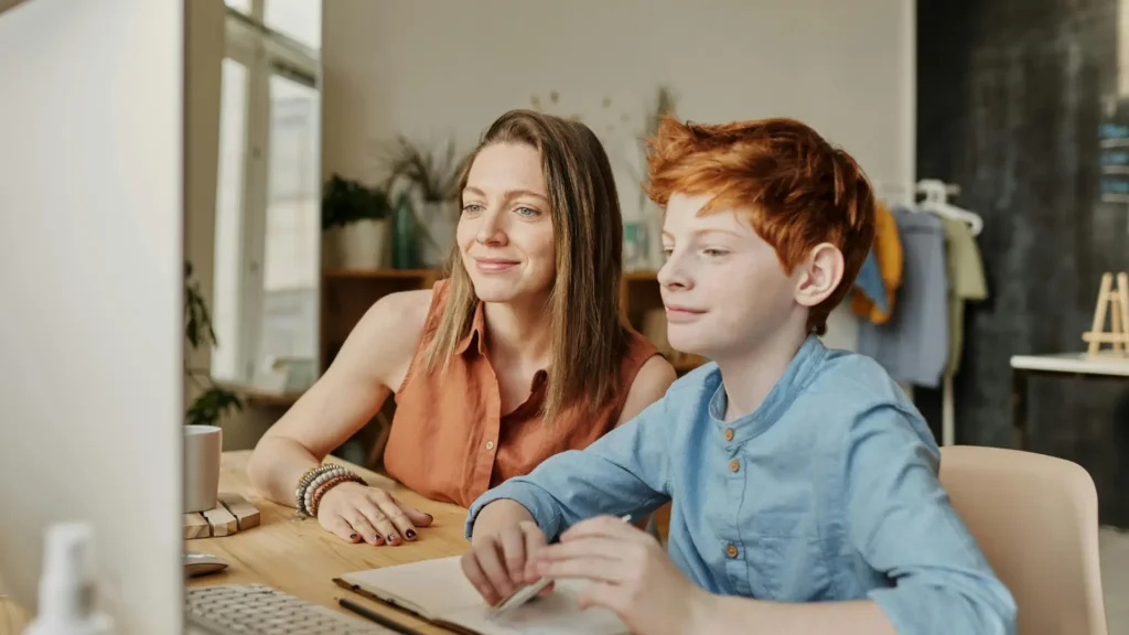 Adult female mother/guardian and young teenaged boy sit at a desk looking at information on screen.