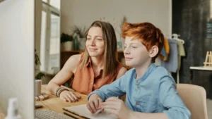 Adult female mother/guardian and young teenaged boy sit at a desk looking at information on screen.