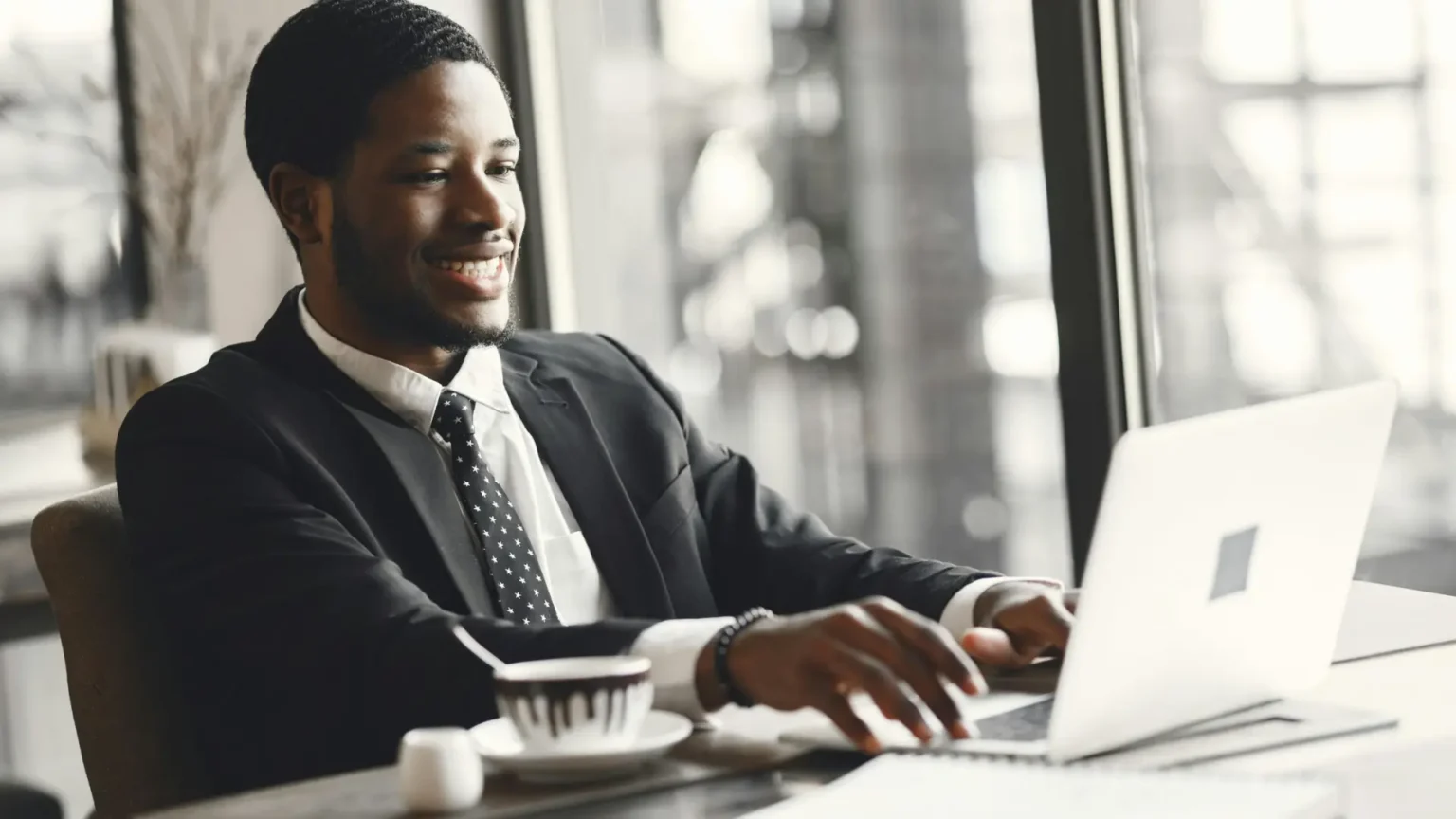 Matering lifelong learning: A smiling professional in a suit works on a laptop at a modern café, with a coffee beside him—reflecting career progression, flexibility, and success through distance learning CMI management qualification.