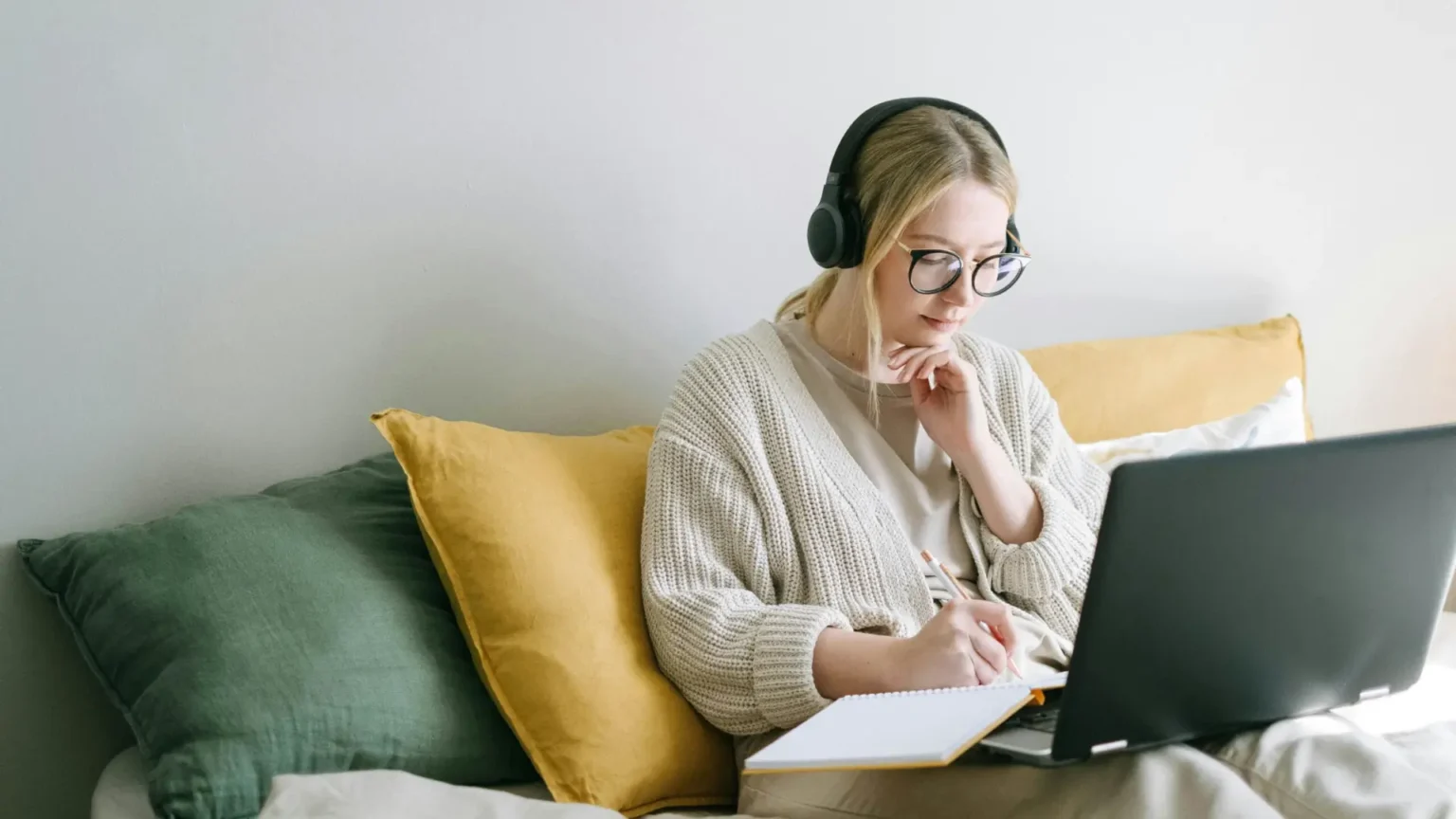 A young woman wearing headphones and glasses sits comfortably on a bed, studying with a laptop and notepad. She appears focused and relaxed, looking at the advantages and disadvantages of studying online.