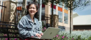 Smiling girl sitting on a bench in the sunshine whilst using her laptop.