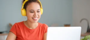 Smiling older teenage student wearing bright yellow headphones looking at her laptop whilst studying at home.