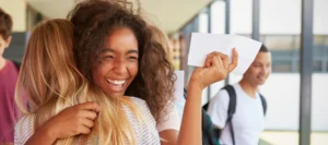 Two teenage girls hugging as one wields her exam results slip with a big smile on her face