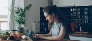 Young woman with curly hair typing on a laptop at a kitchen table while studying for NEC distance learning A levels.
