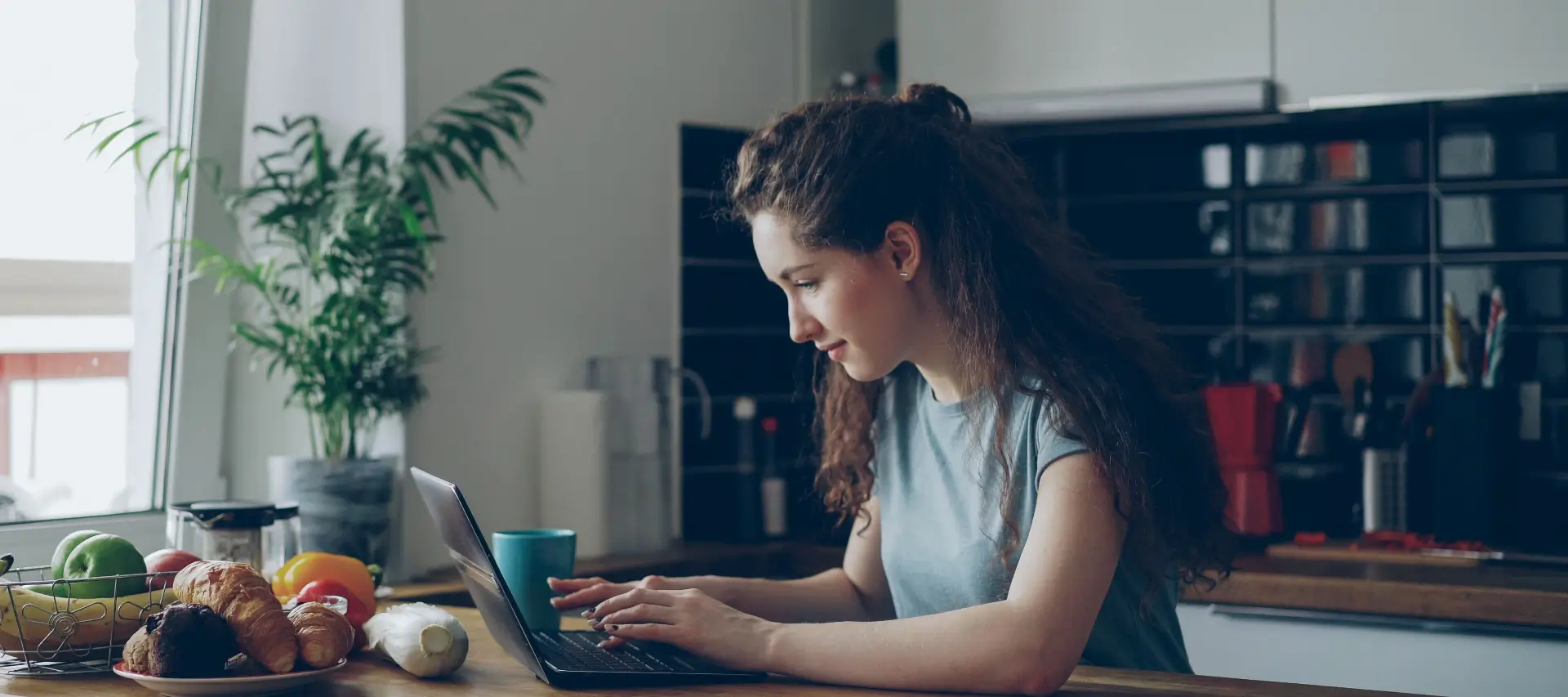 Young woman with curly hair typing on a laptop at a kitchen table while studying for NEC distance learning A levels.