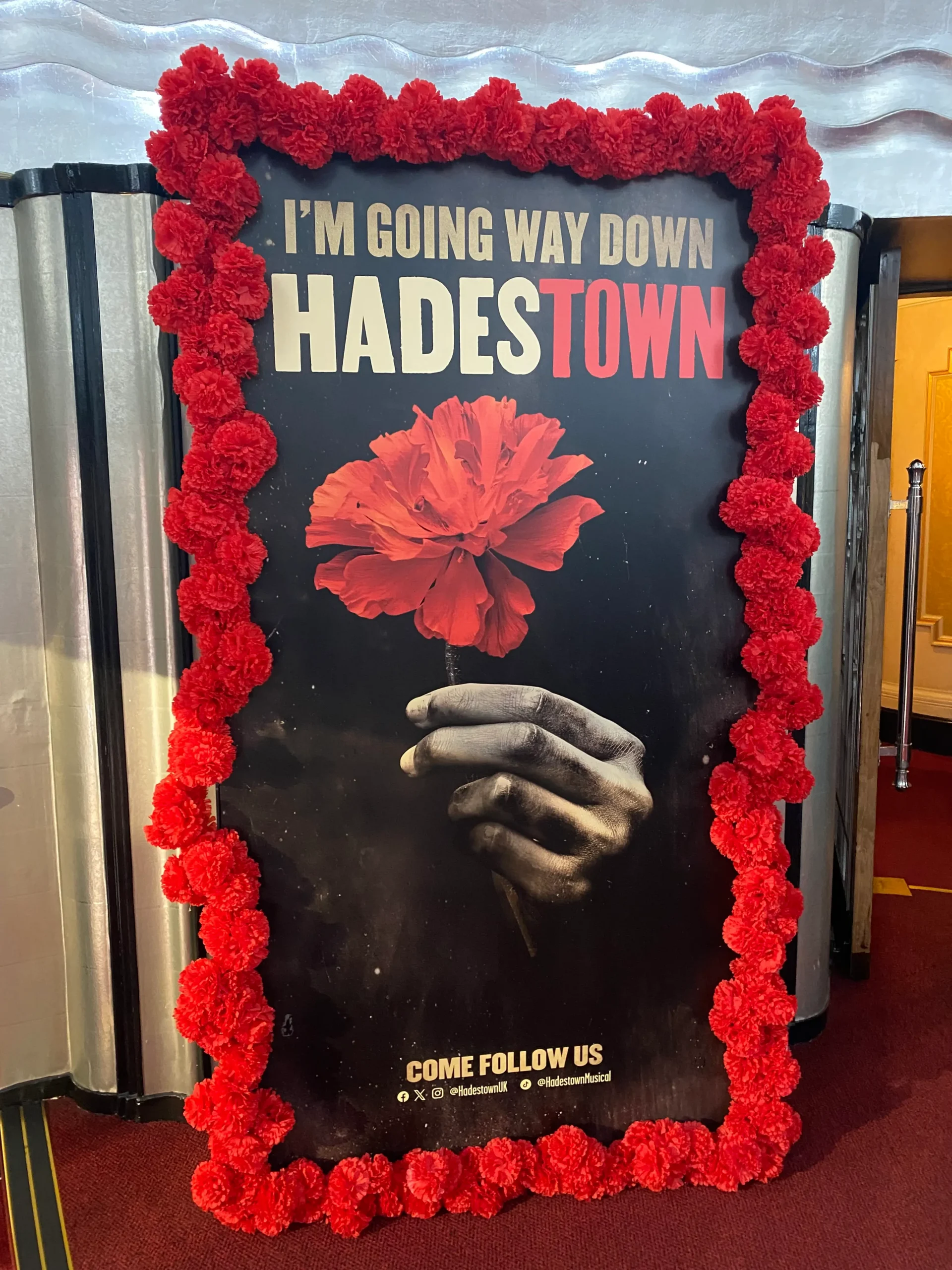 Hadestown poster banner inside the theatre in London. Poster shows a hand holding a red flower on a black background with the words 'Im' going way down, Hadestown'. There is a garland of red flowers all around the poster.